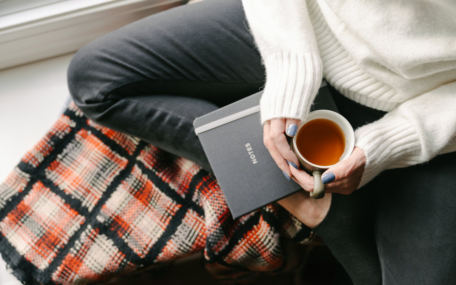 A woman's hands holding a cup of tea and a notebook, wrapped in a plaid blanket