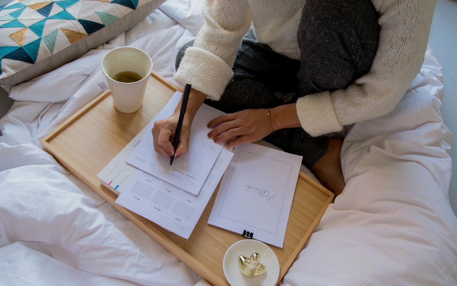 A woman sitting on a bed writing lists on loose pages, with a cup of tea on a wooden tray