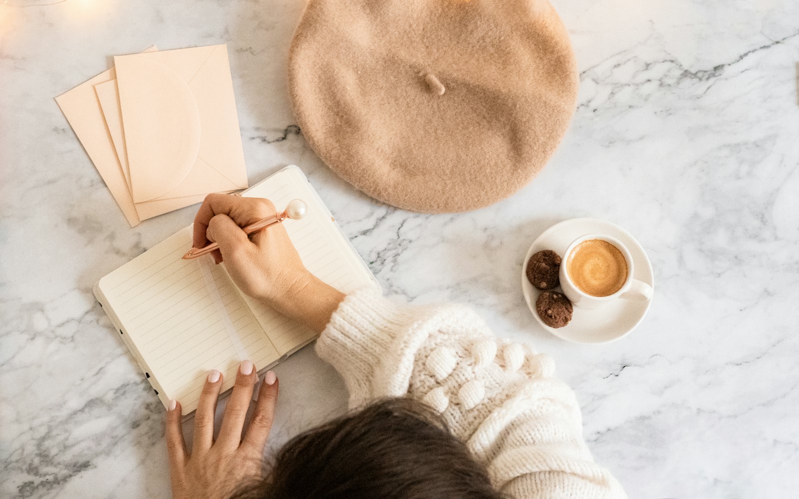 A woman in a cream sweater writing in a notebook next to a cup of coffee