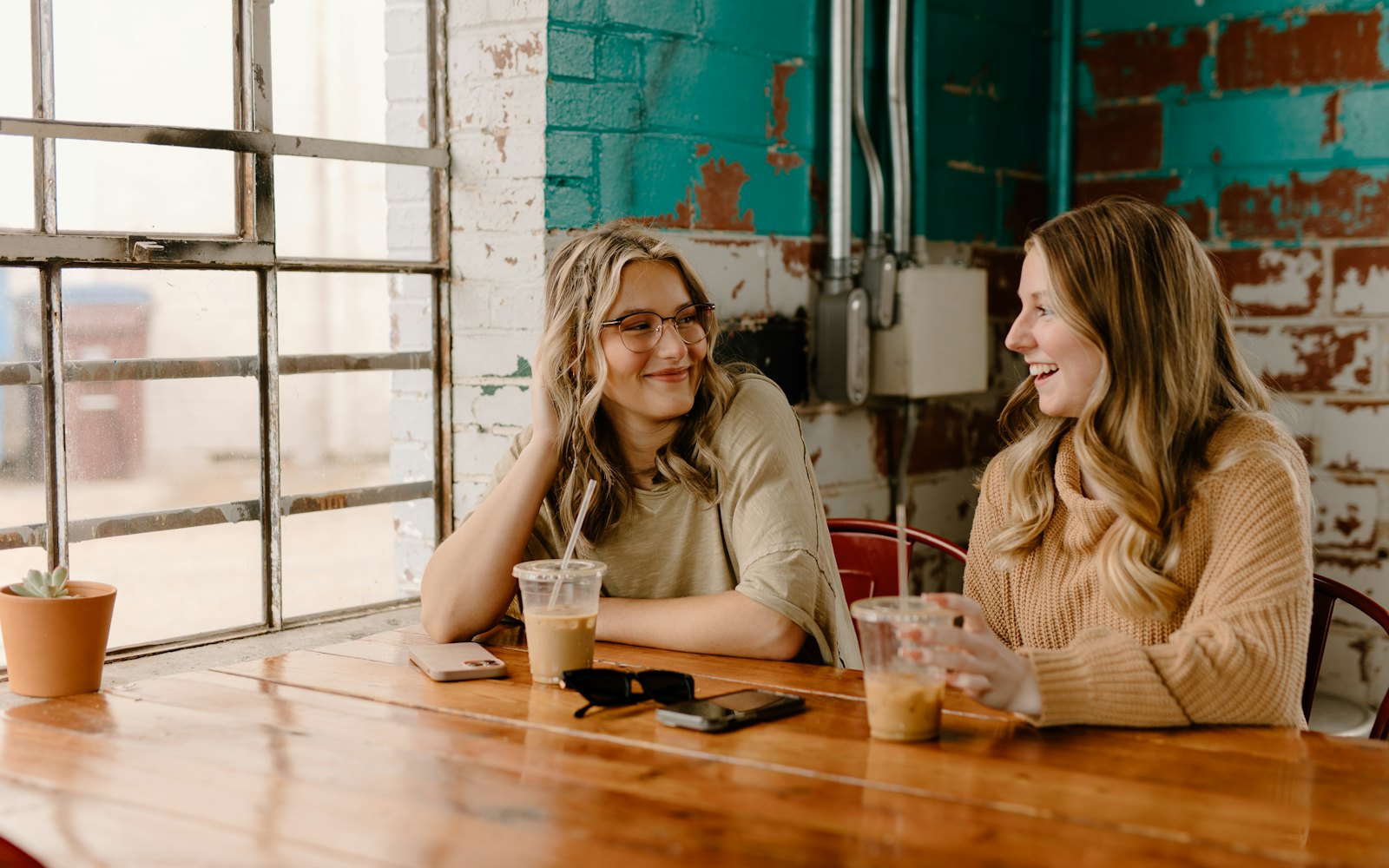 Two women talking over coffee at a wooden table by a big window