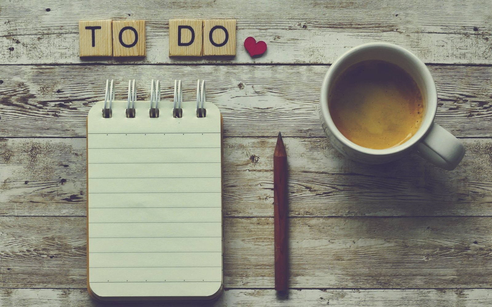 A blank notepad under letter tiles spelling TO DO, next to a cup of tea on a wooden table