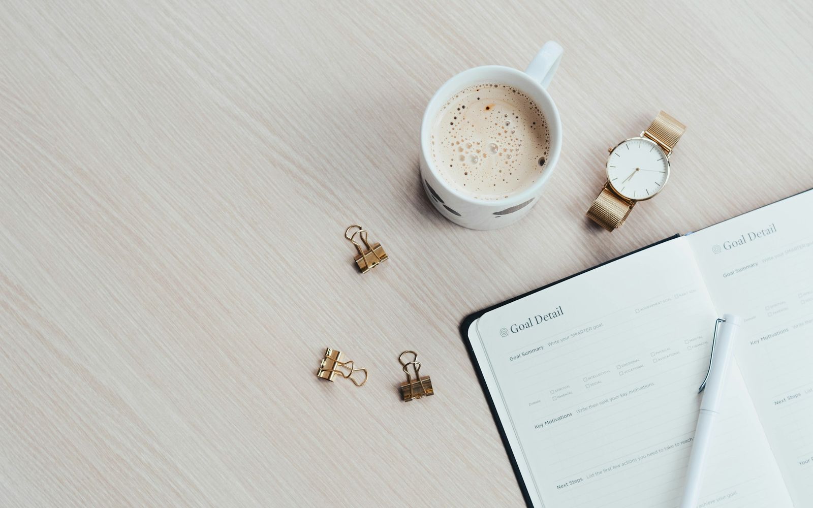An open planner with a pen, a cup of coffee, a wristwatch and paper clips on a light table