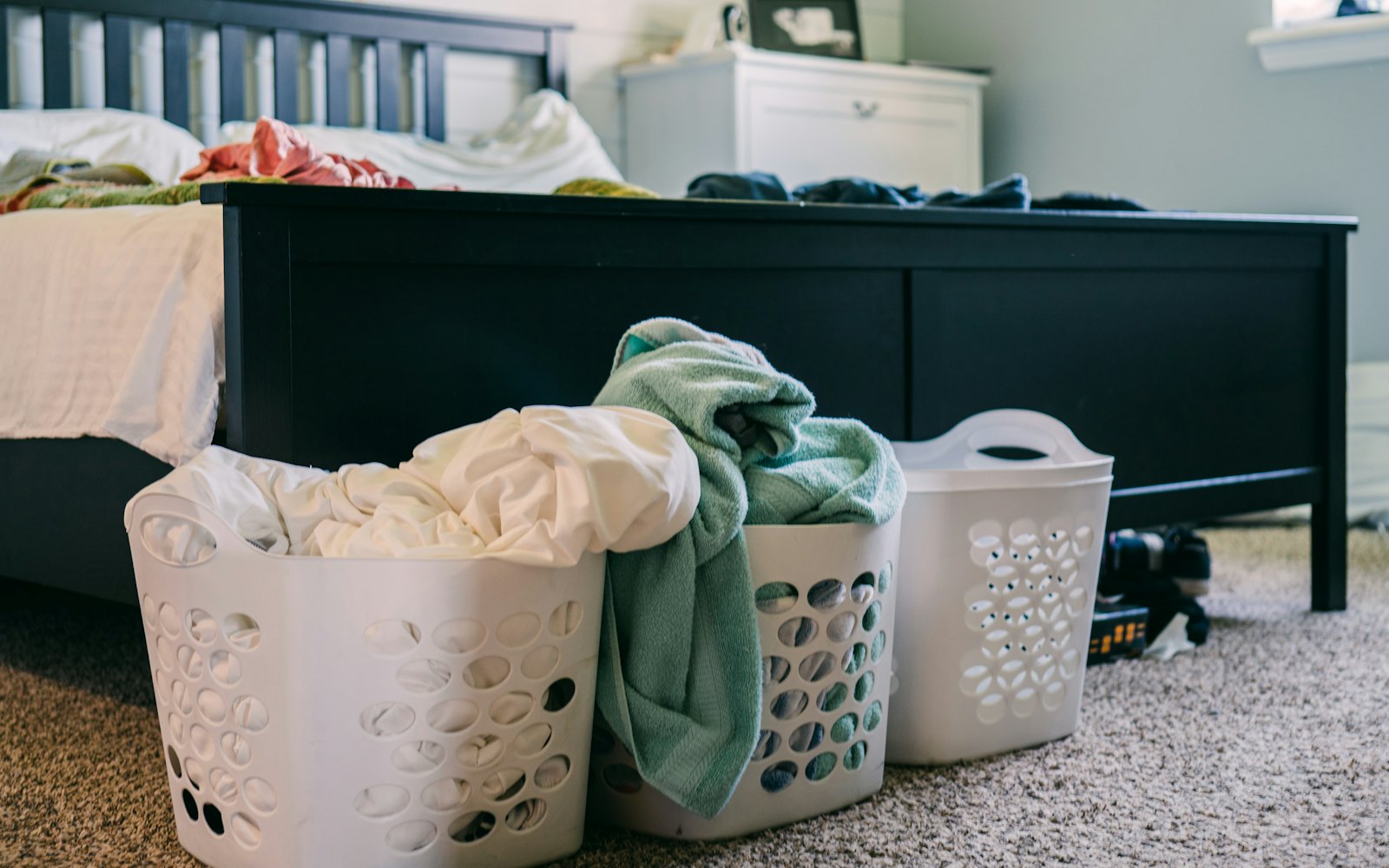 Three laundry baskets full of clothes on the floor beside a bed